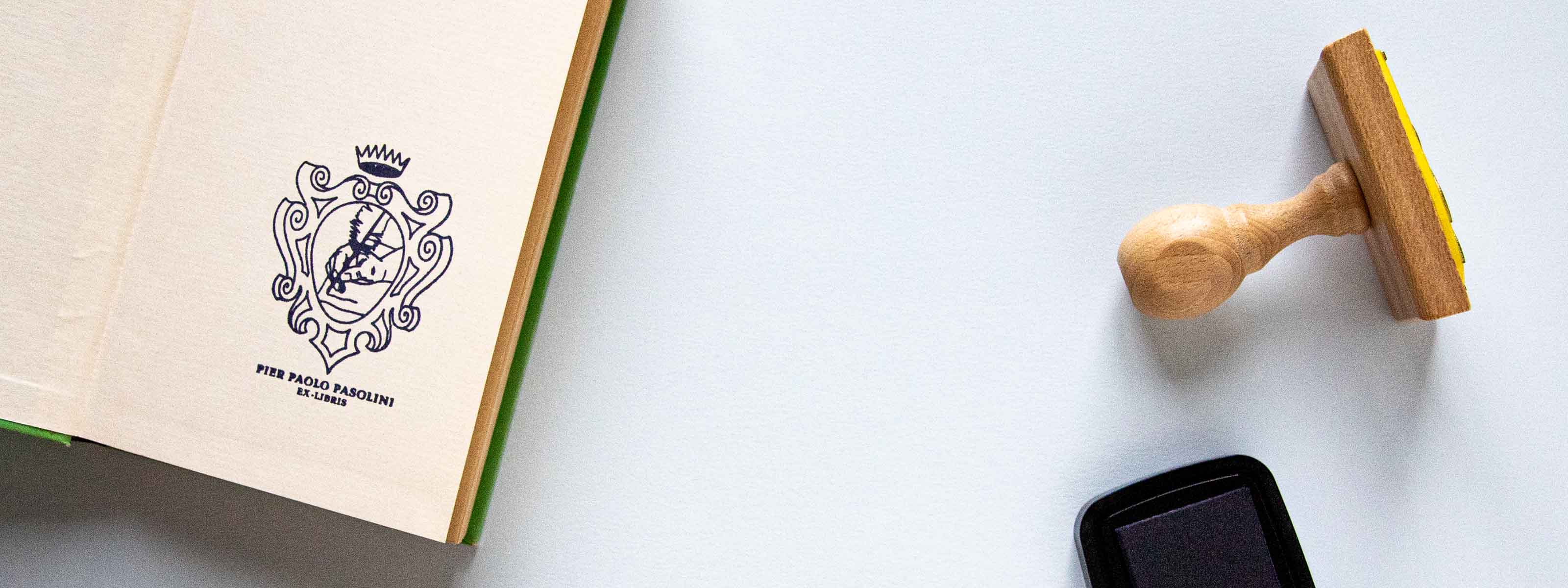 Exlibris personnalisé tamponné sur la page de garde d'un livre ouvert avec un blason héraldique surmonté d’une couronne, à l’intérieur duquel on voit une main écrivant avec une plume