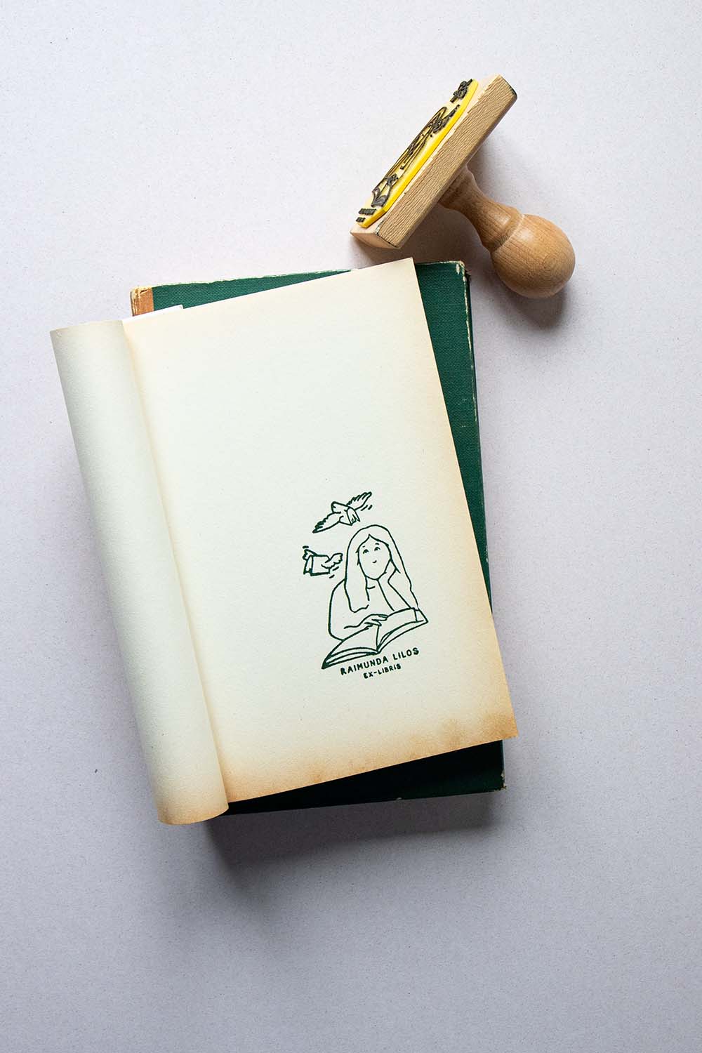 Portadilla de libro estampada con sello exlibris de una mujer leyendo un libro, con dos libros volando sobre ella y sello